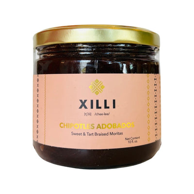A clear jar with a gold lid containing  morita peppers in chipotle adobada sauce. They are sweet and tart. The label is a peach color with gold and black lettering and an image of their logo in gold. 