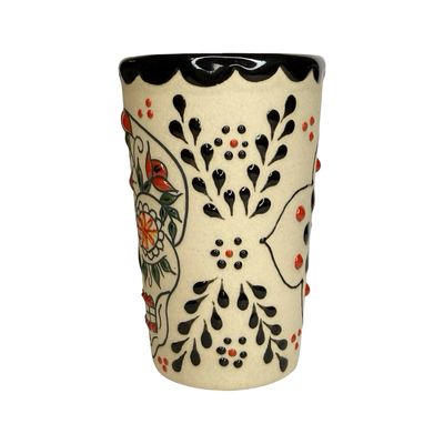 cream ceramic shot glass decorated with colorful sugar skull (Catrina) designs in red, black, and orange.