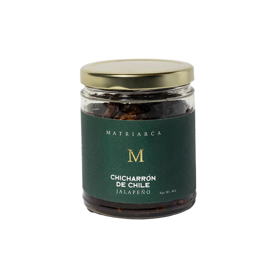 6 oz Glass jar labeled “Chicharrón de Chile Jalapeño” by Matriarca, filled with dark jalapeño pieces in oil, with a deep green label and gold lid.