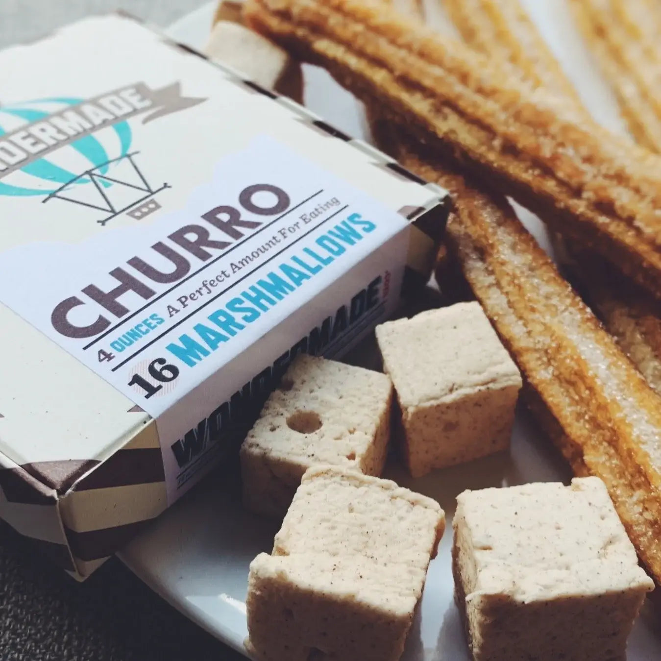 Close-up of cube-shaped churro marshmallows on a plate beside churro sticks and a box labeled “Churro — 16 Marshmallows, 4 ounces.”