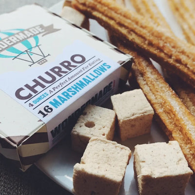 Close-up of cube-shaped churro marshmallows on a plate beside churro sticks and a box labeled “Churro — 16 Marshmallows, 4 ounces.”