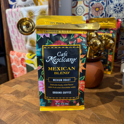 A bag of Café Mexicano Mexican Blend medium roast ground coffee, sealed with a gold coffee scoop clip, displayed on a wooden counter with colorful textiles and additional coffee bags in the background.