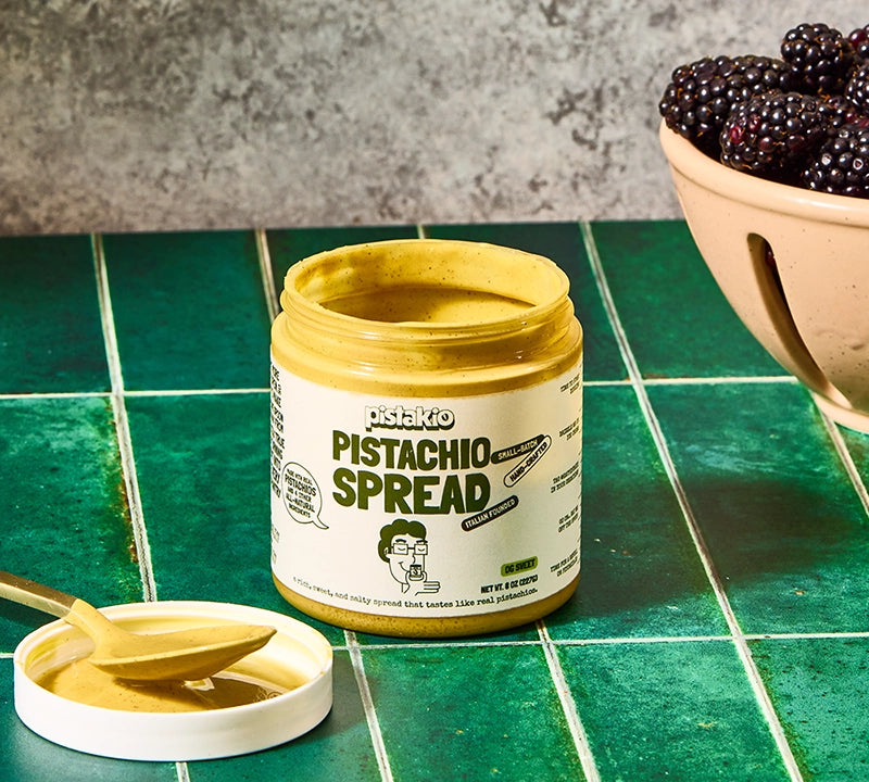 Jar of pistachio spread on a green tiled surface with a bowl of blackberries in the background.