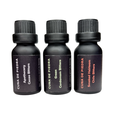 Three small matte black bottles labeled “Cuna de Piedra Bitters” in flavors Apothecary Cacao, Green Cardamom, and Smoked Heirloom Chile, standing side by side.