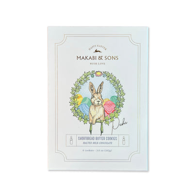 A beautifully designed box of Makabi & Sons Easter Holiday Cookies - Pasha, featuring an illustration of a rabbit surrounded by an Easter wreath with pastel-colored eggs. The packaging highlights shortbread butter cookies with malted milk chocolate, weighing 3.6 oz (102g).