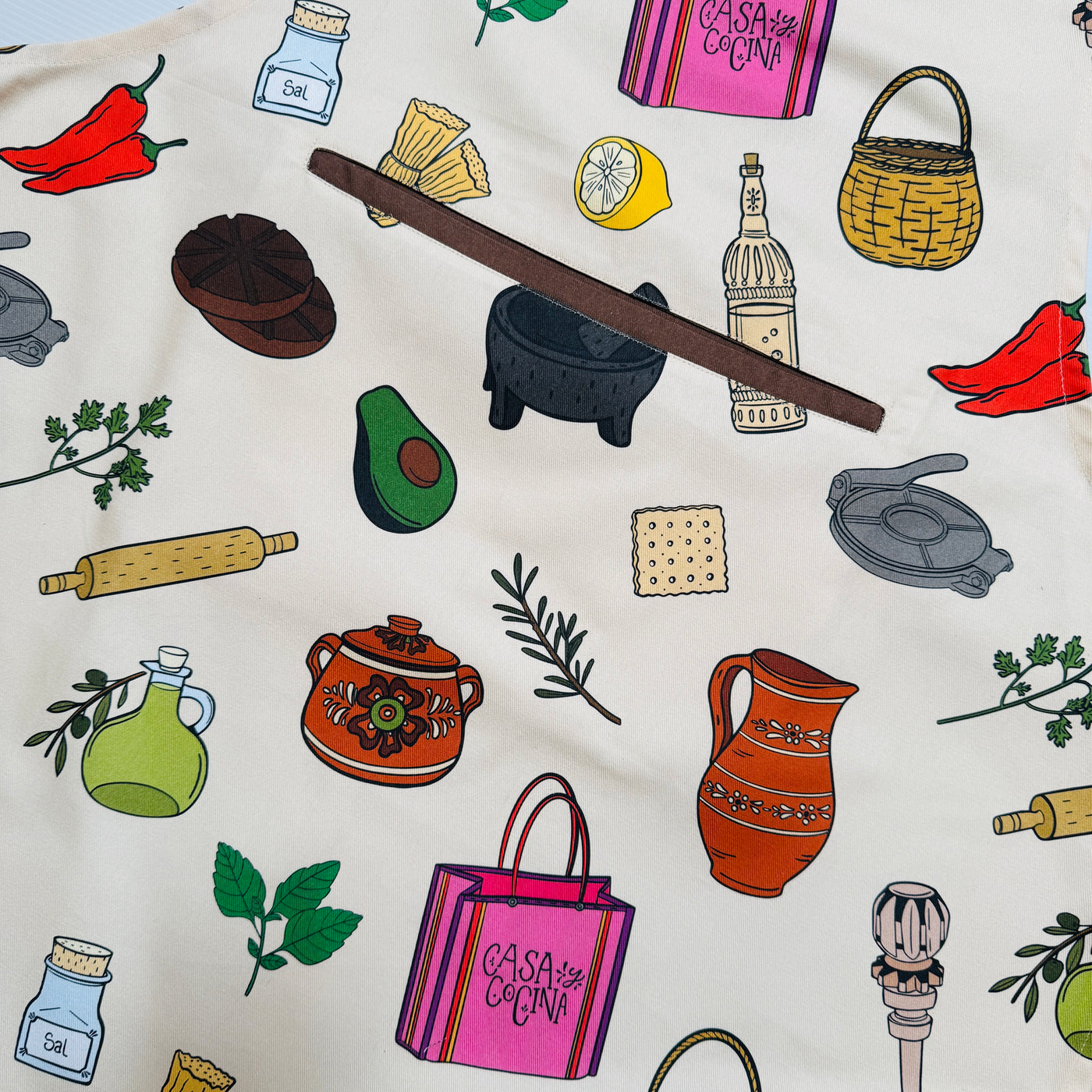 close up view of a Cream apron with colorful illustrated Mexican kitchen icons—molinillo, molcajete, jarras, chiles, avocado—plus a center pocket, adjustable neck, and brown ties.