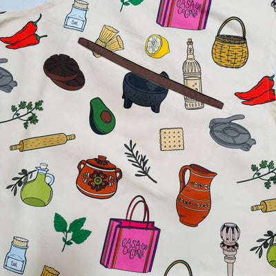 close up view of a Cream apron with colorful illustrated Mexican kitchen icons—molinillo, molcajete, jarras, chiles, avocado—plus a center pocket, adjustable neck, and brown ties.
