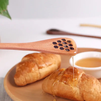 Close-up of a wooden honey spoon with honeycomb-shaped holes, drizzling honey over a pastry on a wooden plate.