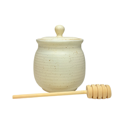 Speckled stoneware honey jar with matching lid and wooden honey dipper laying in front of the jar.