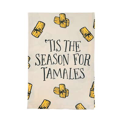 A beige kitchen towel featuring the phrase "'Tis the Season for Tamales" in bold black lettering, surrounded by illustrations of golden tamales.