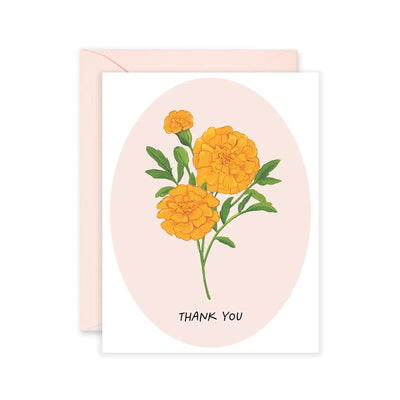 Greeting card with an illustration of three bright orange marigold flowers and green leaves centered on a soft pink oval background, with the words "Thank You" at the bottom. Pale pink envelope included.