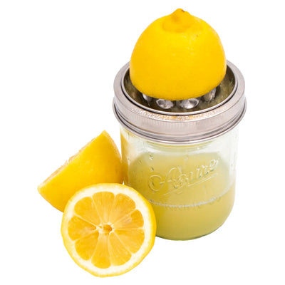 A stainless steel mason jar juicing lid attached to a wide-mouth glass jar, juicing a lemon with fresh juice inside, surrounded by sliced lemons.