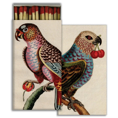  A decorative matchbox featuring a vintage-style illustration of two colorful parrots. One parrot is holding a cherry in its claw, while the other holds two cherries in its beak. The birds are perched on tree branches, and the matchbox lid partially covers the artwork, creating a layered effect.