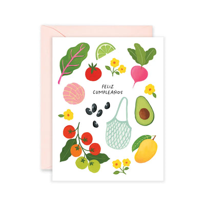 Greeting card with the phrase "Feliz Cumpleaños" surrounded by illustrated fruits, vegetables, flowers, and a market bag, including avocado, mango, lime, tomato, beans, and pan dulce, with a light pink envelope.