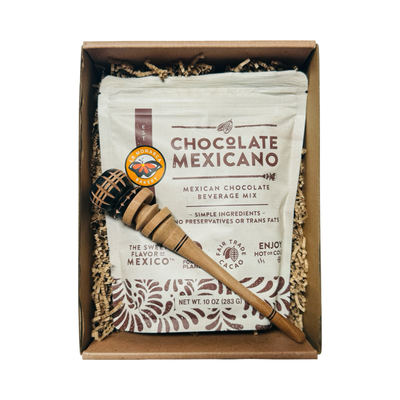 Top view of open box showing a 10 oz pouch of “Chocolate Mexicano” mix and a small wooden molinillo nestled in kraft paper.