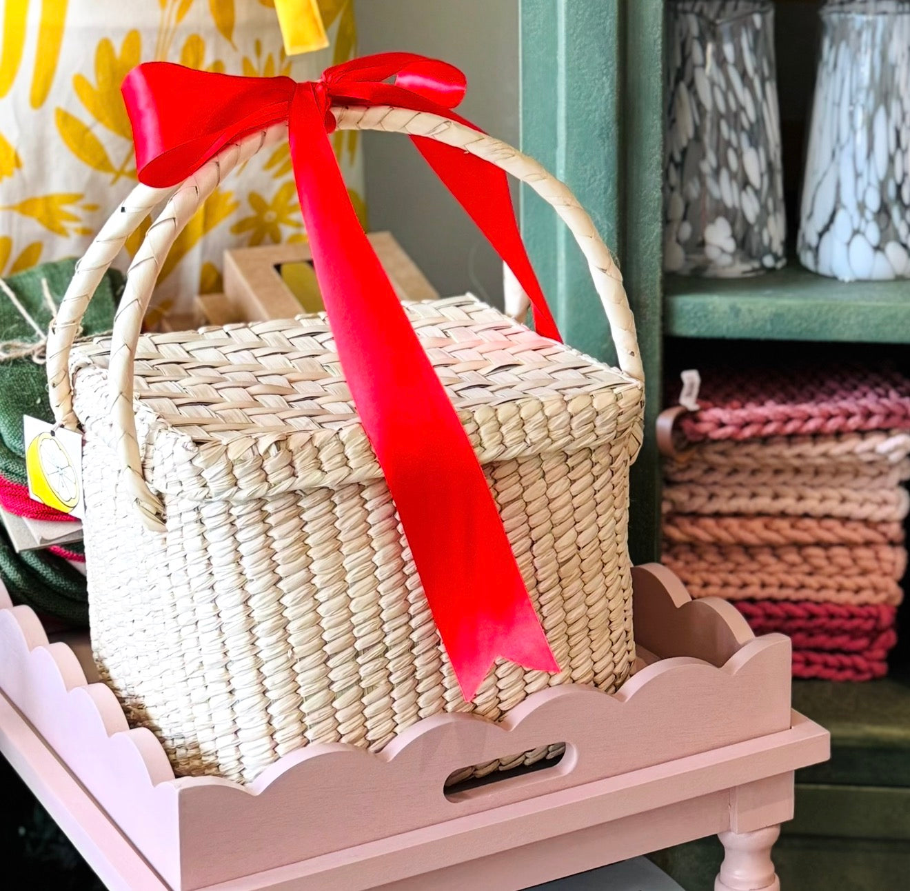 Handwoven palm picnic basket with lid and double handles tied with a red ribbon sitting on a pink bar cart.
