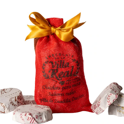 A festive red mesh gift bag tied with a shiny yellow satin ribbon, labeled "Villa Real." Surrounding the bag are individually wrapped chocolate discs in white paper with red and gold lettering. The set includes assorted Mexican hot chocolate flavors.