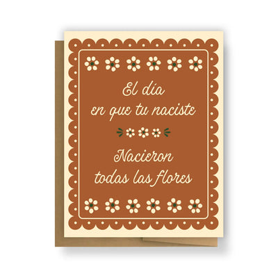 A greeting card with a warm brown background and cream-colored decorative border featuring small flowers. The text reads "El día en que tú naciste, nacieron todas las flores", inspired by the traditional Mexican song Las Mañanitas. The card is paired with a kraft envelope.