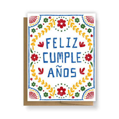 A festive greeting card with the words "Feliz Cumpleaños" in bold blue letters, surrounded by a colorful floral folk-art design in red, yellow, green, and blue. The card is paired with a kraft envelope.