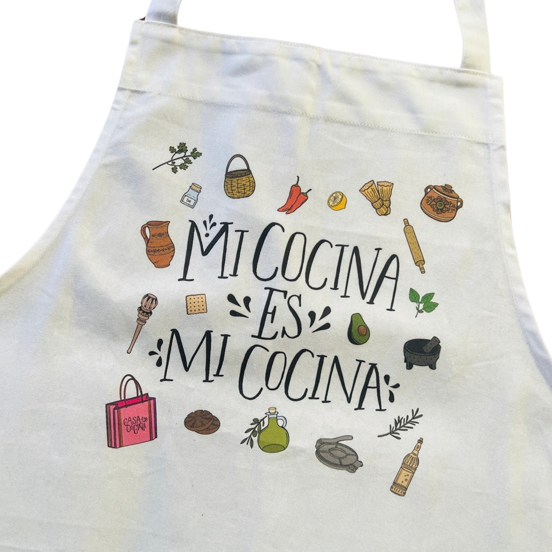 Close up view of a white apron with the phrase Mi Cocina es Mi Cocina in black lettering featurng various images of Mexican kitchen essentials such as tortilla press, pitcher, molcajete and pots.