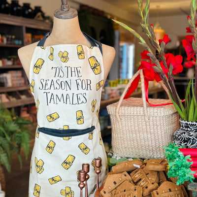 This image features a festive apron with a light beige background and a repeating pattern of tamales. The apron includes the playful text "Tis the Season for Tamales" at the top with black neck and waist ties.
