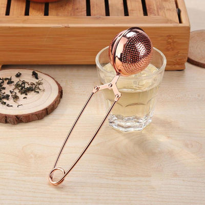 Rose gold colored metal tea steeper sitting on tpo of clear glass in kitchen setting