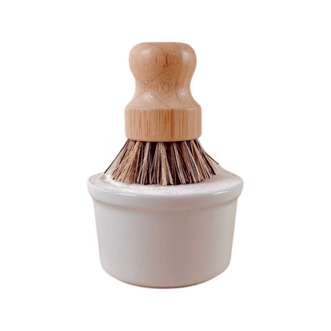 Dishwashing brush with wood handle sitting on top of white cylindrical soap bowl from previous photo