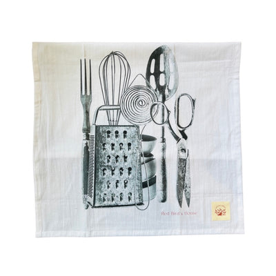 White kitchen towel with silver utensils like a whisk, slotted spoon, scissors, fork and whisk. folded in half.