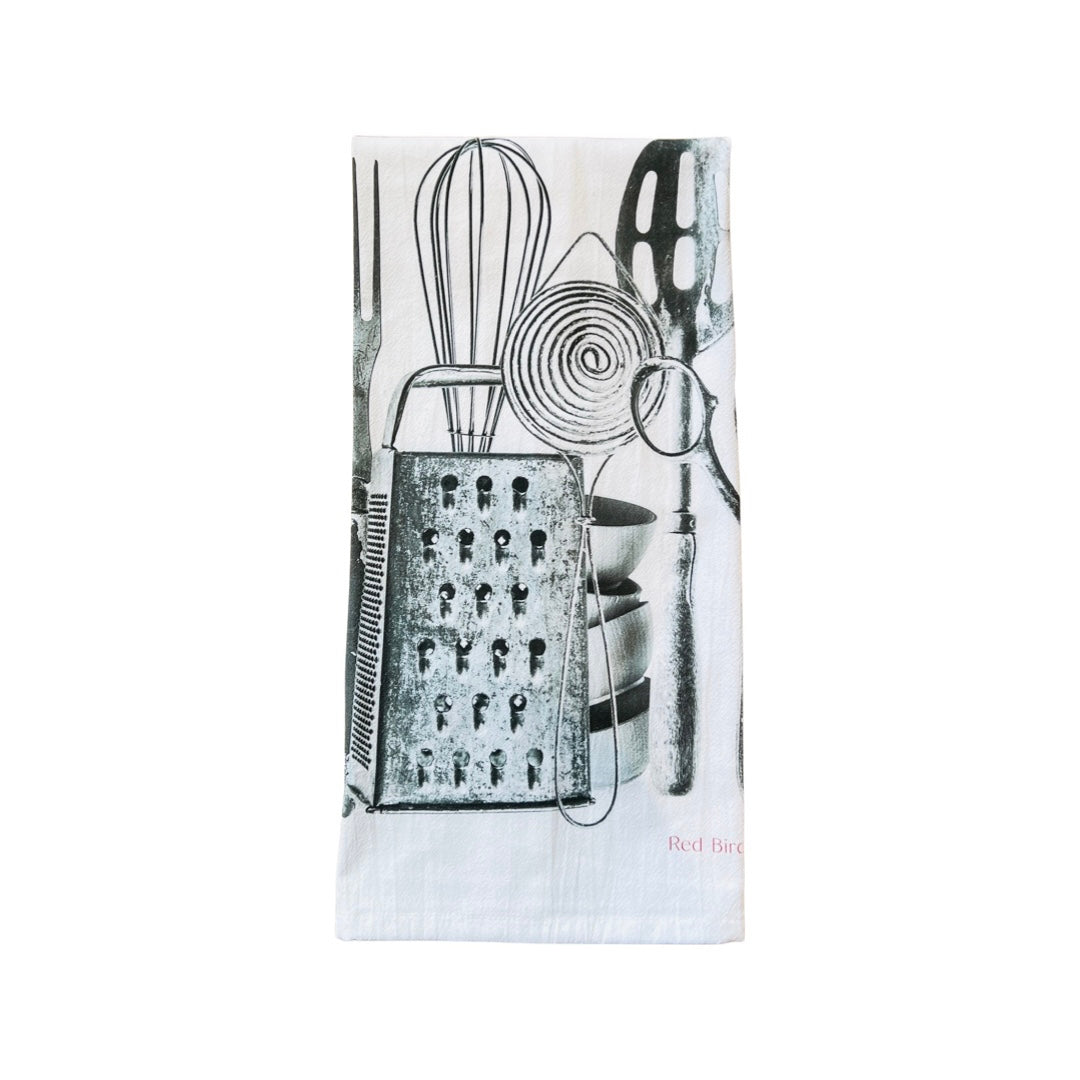 White kitchen towel with silver utensils like a whisk, slotted spoon and whisk. folded in quarters.